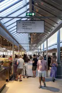 Busy indoor market corridor with directional signage pointing to a food hall, ATM, metro, and restrooms