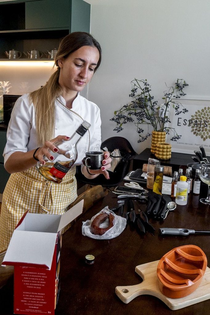A chef pouring brandy from a bottle into a small metal cup at a kitchen prep table