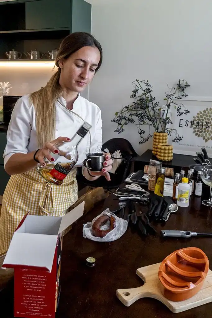 A chef pouring brandy from a bottle into a small metal cup at a kitchen prep table