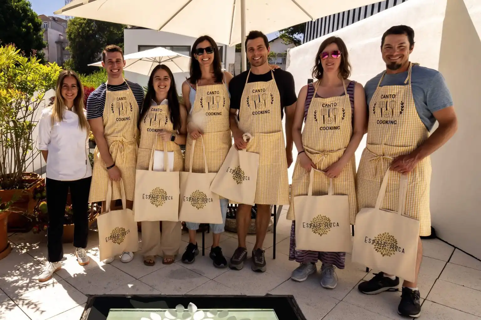 A group of cooking class guests wearing yellow gingham aprons and holding tote bags on a sunny terrace
