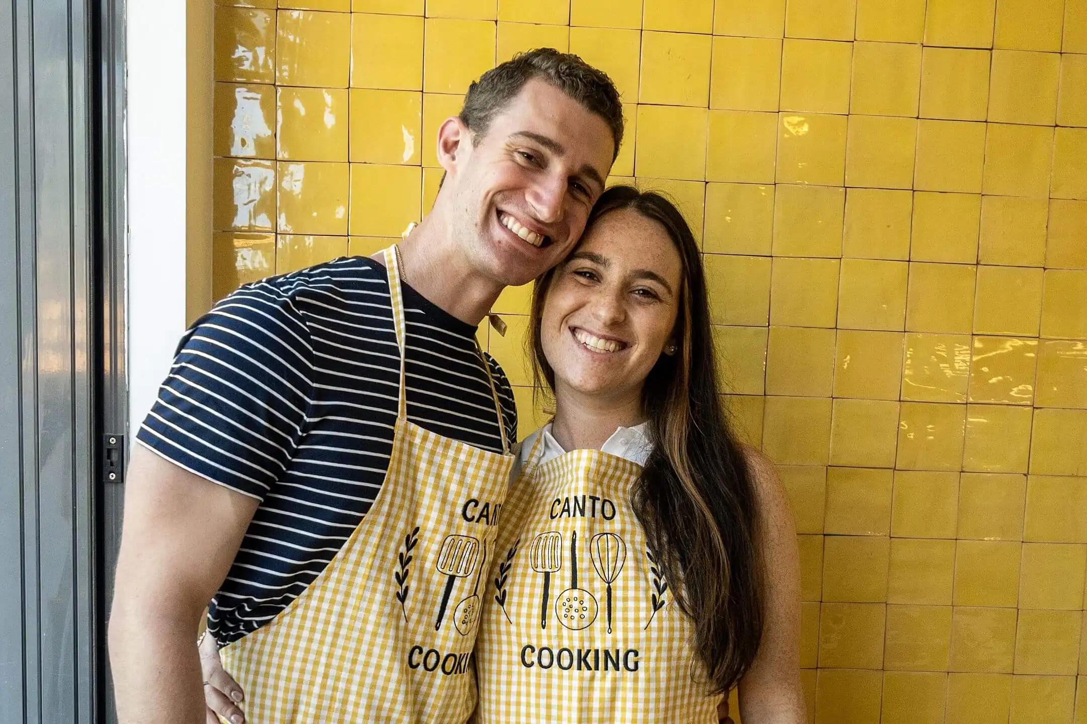 Two Canto Cooking guests in matching yellow gingham aprons smiling against a yellow azulejo wall in Porto