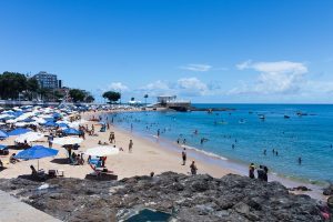 Crowded sandy beach with blue umbrellas and a white historic fort on a rocky promontory