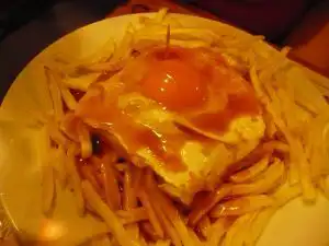 Francesinha sandwich topped with a fried egg and tomato-beer sauce, served on a bed of thin fries
